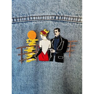 May include: Embroidered patch depicting a couple on a balcony with a sunset. The woman wears a red dress and the man a black coat. The background is a light blue denim.