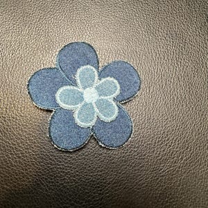 May include: A denim flower patch with a five-petal design. The petals are a dark blue denim, and the center petals are a lighter blue denim. The patch has a white stitched border.