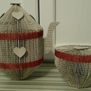 Pode incluir: Um conjunto de chá decorativo composto por uma chaleira e uma chávena, feito de páginas de livros dobradas. A chaleira tem uma pega, um bico e dois enfeites em forma de coração. Ambas as peças são adornadas com uma fita vermelha. Um artigo de decoração para casa único e artesanal.