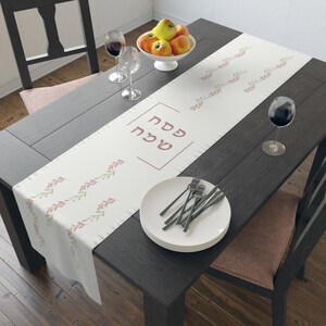May include: A white table runner with a pink and green leafy pattern and the Hebrew text "Happy Passover" in pink. The runner is on a dark wood table with a bowl of apples, two wine glasses, and a stack of white plates.