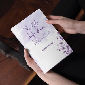 Può includere: Un diario bianco con uno sfondo acquerellato viola e il testo "Thank You Hashem Daily Gratitude Journal" e "Family Friedman". Il diario ha un design floreale con fiori e foglie viola.