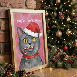 Op de afbeelding: Een ingelijste print met een grijze kat met een kerstmuts en een rode halsband met belletjes, met een zuurstok. De roze achtergrond heeft sneeuwvlokken en de tekst "Santa's favorite Meow". Kerstversiering.