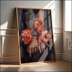Pode incluir: Obra de arte emoldurada com uma pintura detalhada de mãos entrelaçadas com fio vermelho, segurando três grandes peónias cor de coral. A obra de arte é definida contra um fundo escuro e texturizado e emoldurada em madeira clara.