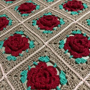 Könnte beinhalten: Eine gehäkelte Decke mit einem sich wiederholenden Muster aus roten Rosen und grünen Blättern. Die Decke ist aus beigefarbenem Garn gefertigt und hat einen rosa Rand.