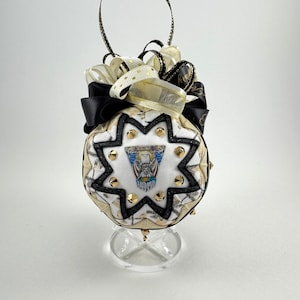 May include: A decorative ornament with a starburst design, featuring a central emblem and gold-colored studs. The ornament is adorned with black, gold, and white ribbons and has a loop for hanging. It is displayed on a clear stand.