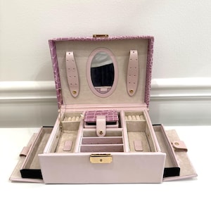 May include: A light purple jewelry box with a crocodile-embossed exterior and a gold-tone metal latch. The interior features a beige velvet lining, an oval mirror, two side earring tabs, a central lidded compartment, and multiple storage slots.