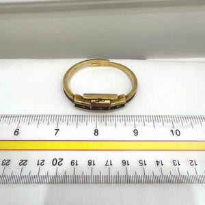 May include: A gold-coloured bracelet with a rectangular clasp and black accents. The bracelet is approximately 18 cm in circumference. The clasp features small cross-shaped designs. The bracelet is on a white surface with a ruler.