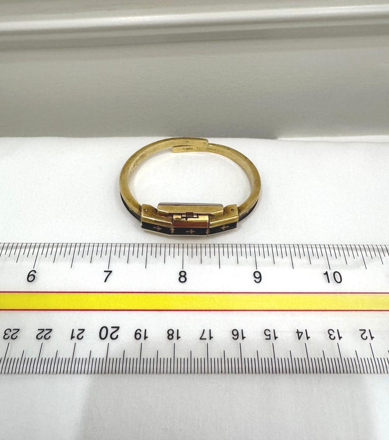 May include: A gold-coloured bracelet with a rectangular clasp and black accents. The bracelet is approximately 18 cm in circumference. The clasp features small cross-shaped designs. The bracelet is on a white surface with a ruler.