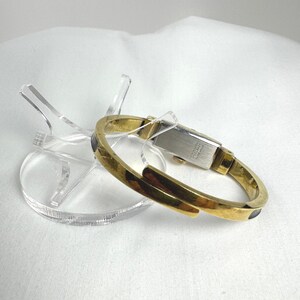 May include: A gold-coloured bracelet watch with a rectangular face and a hinged design. The watch features a gold band with dark accents and rests on a clear acrylic display stand. The watch is a vintage style.