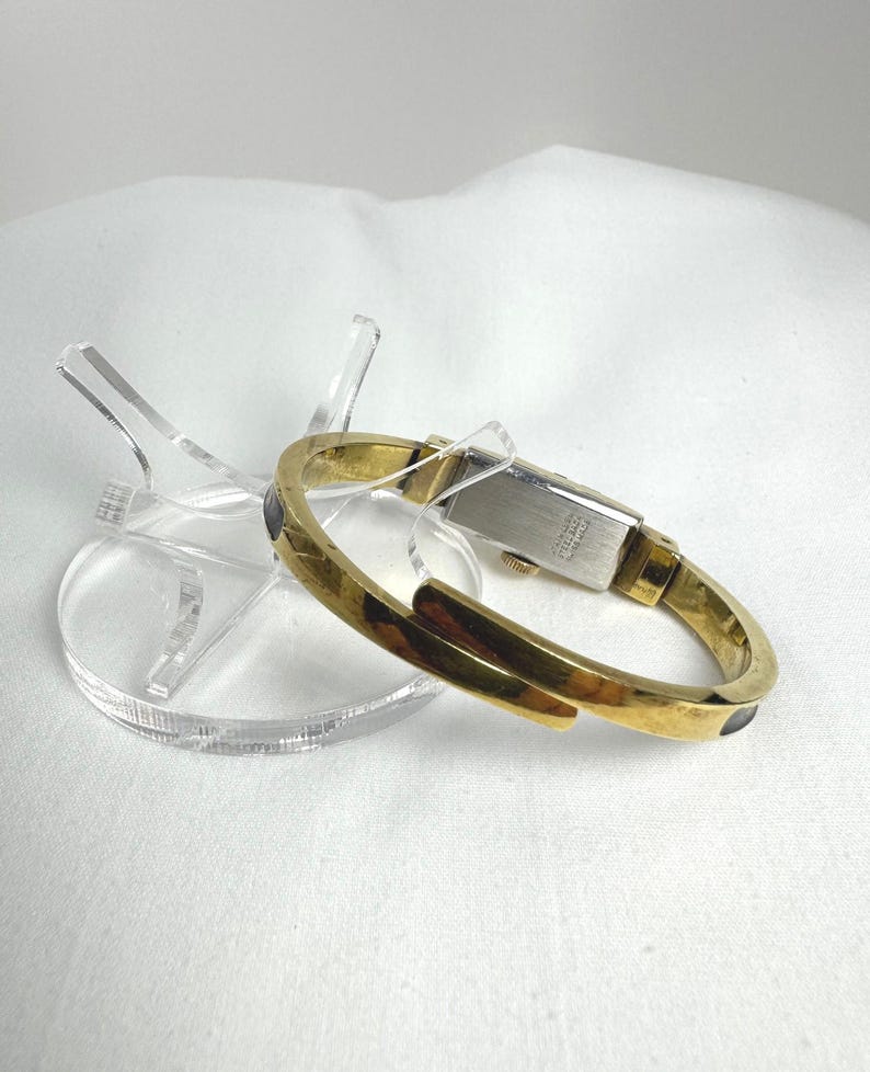 May include: A gold-coloured bracelet watch with a rectangular face and a hinged design. The watch features a gold band with dark accents and rests on a clear acrylic display stand. The watch is a vintage style.
