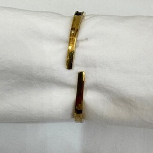 May include: A gold-coloured cuff bracelet with an open design. The bracelet has a slightly curved shape and a polished finish, reflecting light. The ends of the bracelet are flat and slightly tapered. The jewellery is displayed on a white surface.