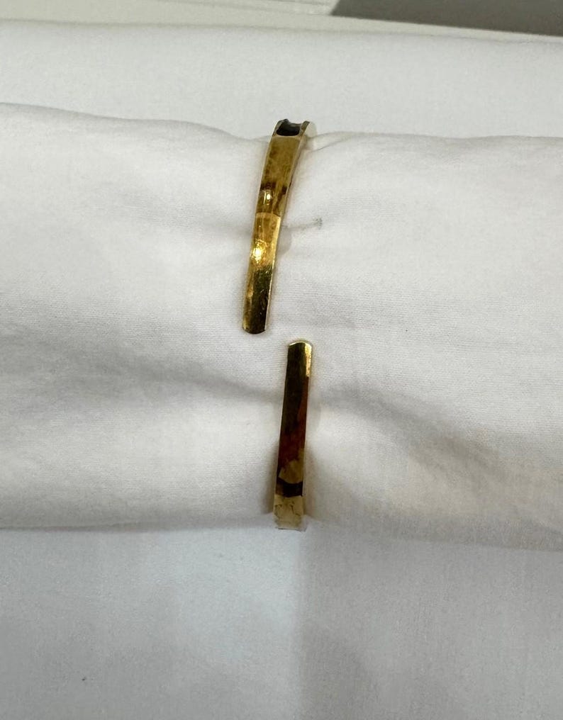 May include: A gold-coloured cuff bracelet with an open design. The bracelet has a slightly curved shape and a polished finish, reflecting light. The ends of the bracelet are flat and slightly tapered. The jewellery is displayed on a white surface.