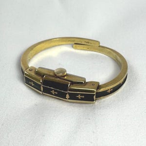 May include: A gold-coloured bracelet watch with a rectangular black face and a gold crown. The black face is decorated with gold fleur-de-lis symbols. The bracelet is a hinged design.