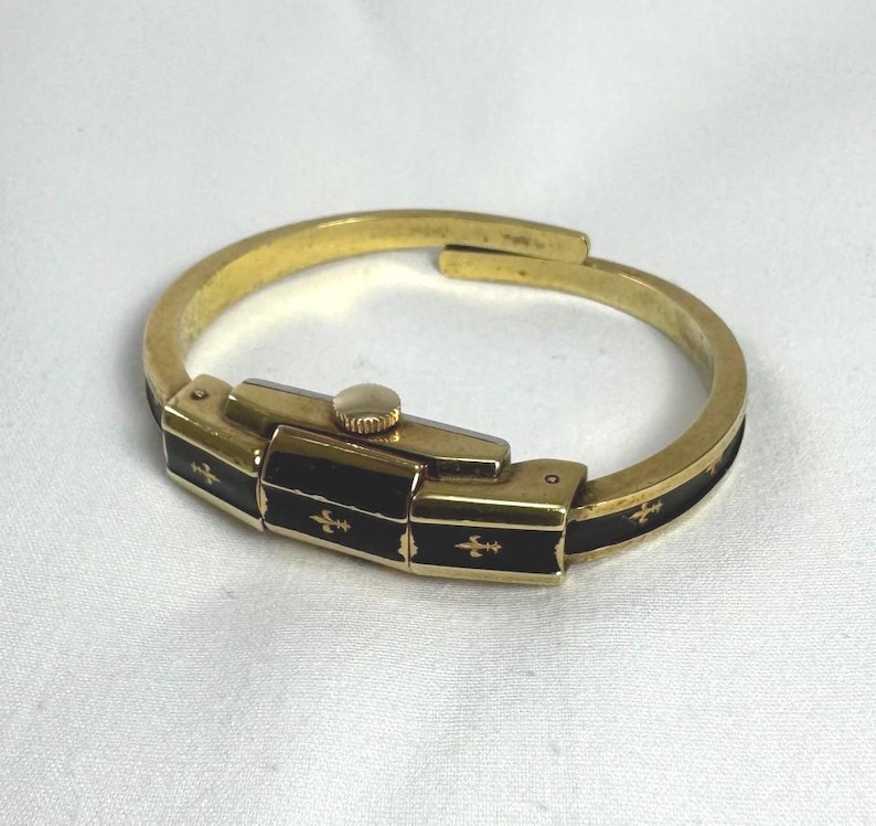 May include: A gold-coloured bracelet watch with a rectangular black face and a gold crown. The black face is decorated with gold fleur-de-lis symbols. The bracelet is a hinged design.