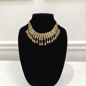 May include: A gold-toned statement necklace featuring a series of curved, textured metal plates arranged in a semi-circular collar design. Small, conical bell-shaped charms hang from the bottom edge of each plate, creating a layered, fringe-like effect.