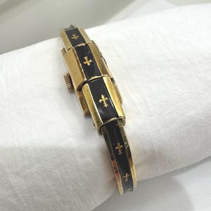 May include: A gold watch with black enamel sections and fleur-de-lis motifs. The watch has a rectangular, articulated strap and a clasp. The watch is shown on a white cloth.