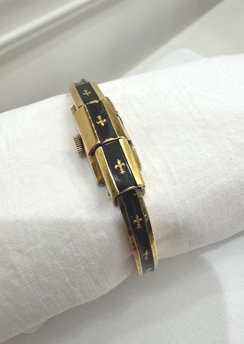 May include: A gold watch with black enamel sections and fleur-de-lis motifs. The watch has a rectangular, articulated strap and a clasp. The watch is shown on a white cloth.