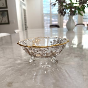 May include: A clear glass bowl with a gold rim and gold floral design. The bowl has three small feet and sits on a reflective surface. The bowl is likely for decorative purposes.