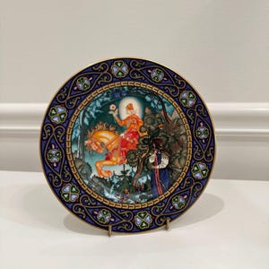 May include: Decorative plate with a detailed illustration of a figure on horseback, set against a forest backdrop. The plate features a blue border with a gold rim and ornate clover-like designs. The artwork is rich in color and detail.