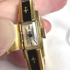 May include: A vintage gold-tone watch with a rectangular face, black enamel accents, and fleur-de-lis details. The watch face displays the brand name "Helios" and "17 Jewels Incabloc". The watch is a hinged bracelet style.