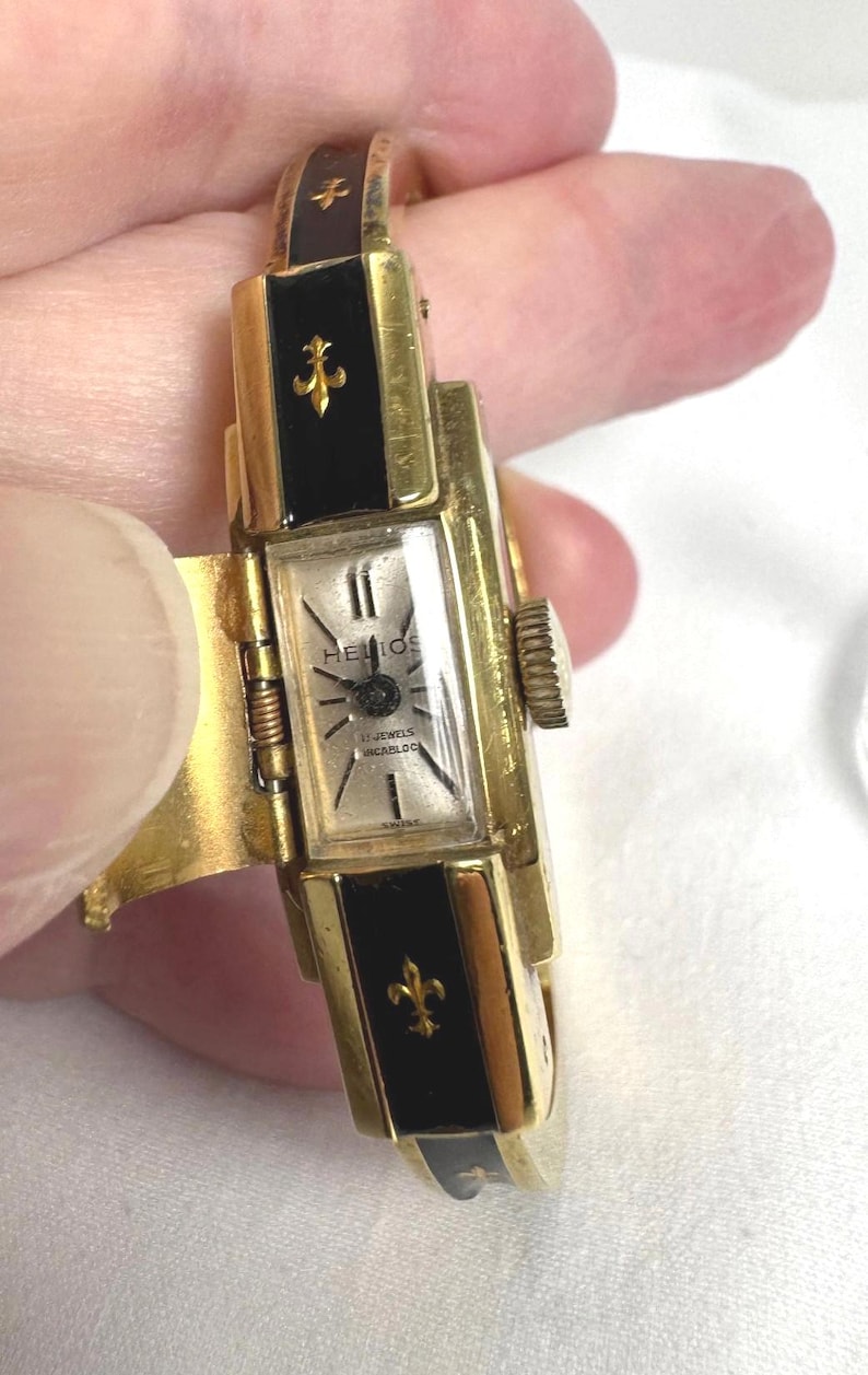 May include: A vintage gold-tone watch with a rectangular face, black enamel accents, and fleur-de-lis details. The watch face displays the brand name "Helios" and "17 Jewels Incabloc". The watch is a hinged bracelet style.