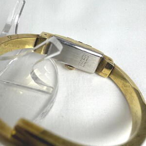 May include: A gold-coloured bangle watch with a rectangular silver watch face. The watch face has the text "Stainless Steel Swiss Made". The watch band is a thick, curved design.