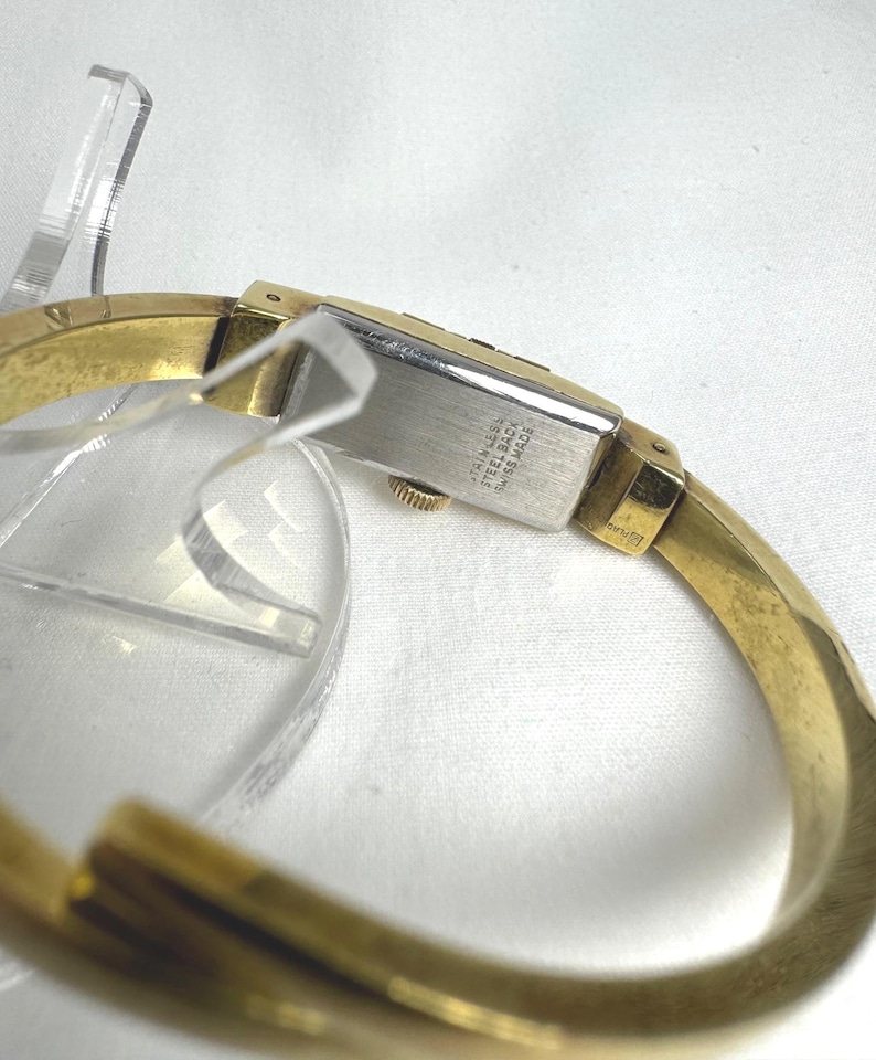 May include: A gold-coloured bangle watch with a rectangular silver watch face. The watch face has the text "Stainless Steel Swiss Made". The watch band is a thick, curved design.