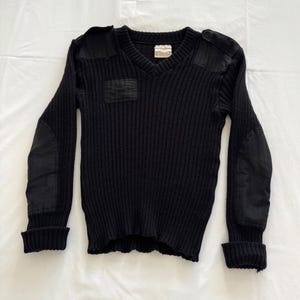 May include: Black V-neck sweater with ribbed knit texture. Features reinforced patches on the shoulders, elbows, and chest. The cuffs and hem are ribbed. A label is visible near the neckline.