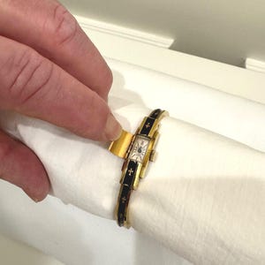 May include: A gold-coloured vintage wristwatch with a rectangular face and black enamel detailing. The watch strap has gold crosses. The watch is held on a white surface.