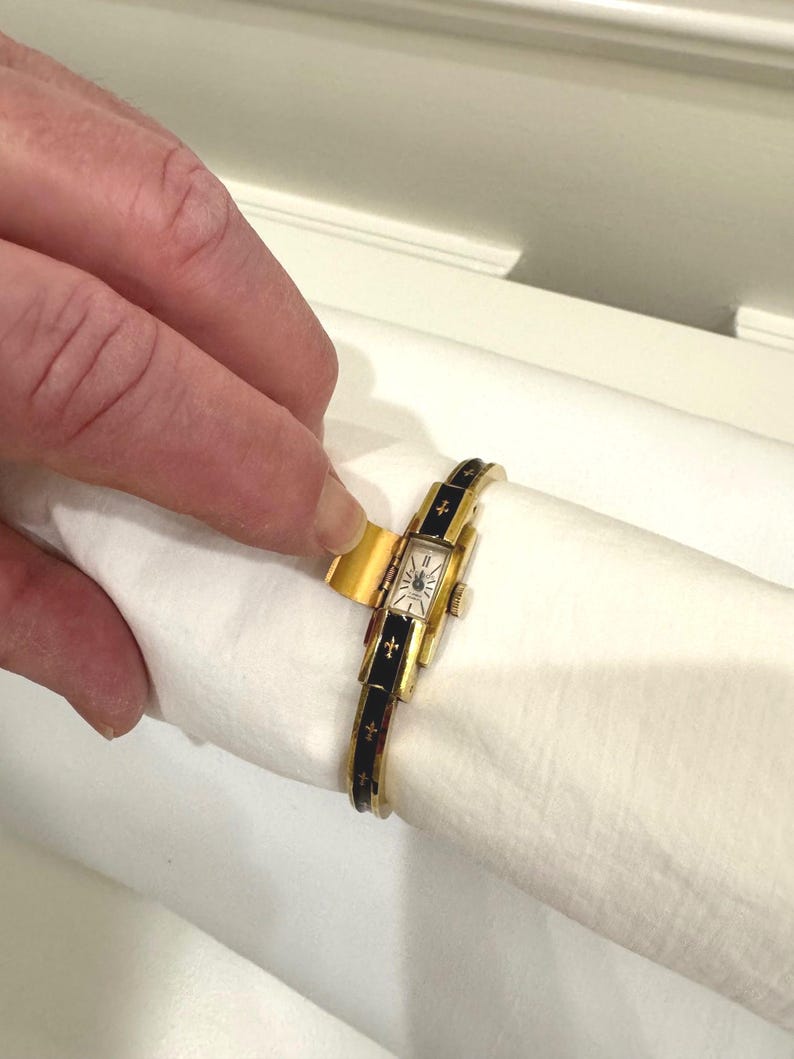 May include: A gold-coloured vintage wristwatch with a rectangular face and black enamel detailing. The watch strap has gold crosses. The watch is held on a white surface.