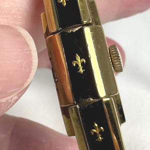 May include: Close-up of a vintage gold-toned watch bracelet with black enamel panels. Each panel features a gold fleur-de-lis emblem. The watch has a textured crown and a classic design.