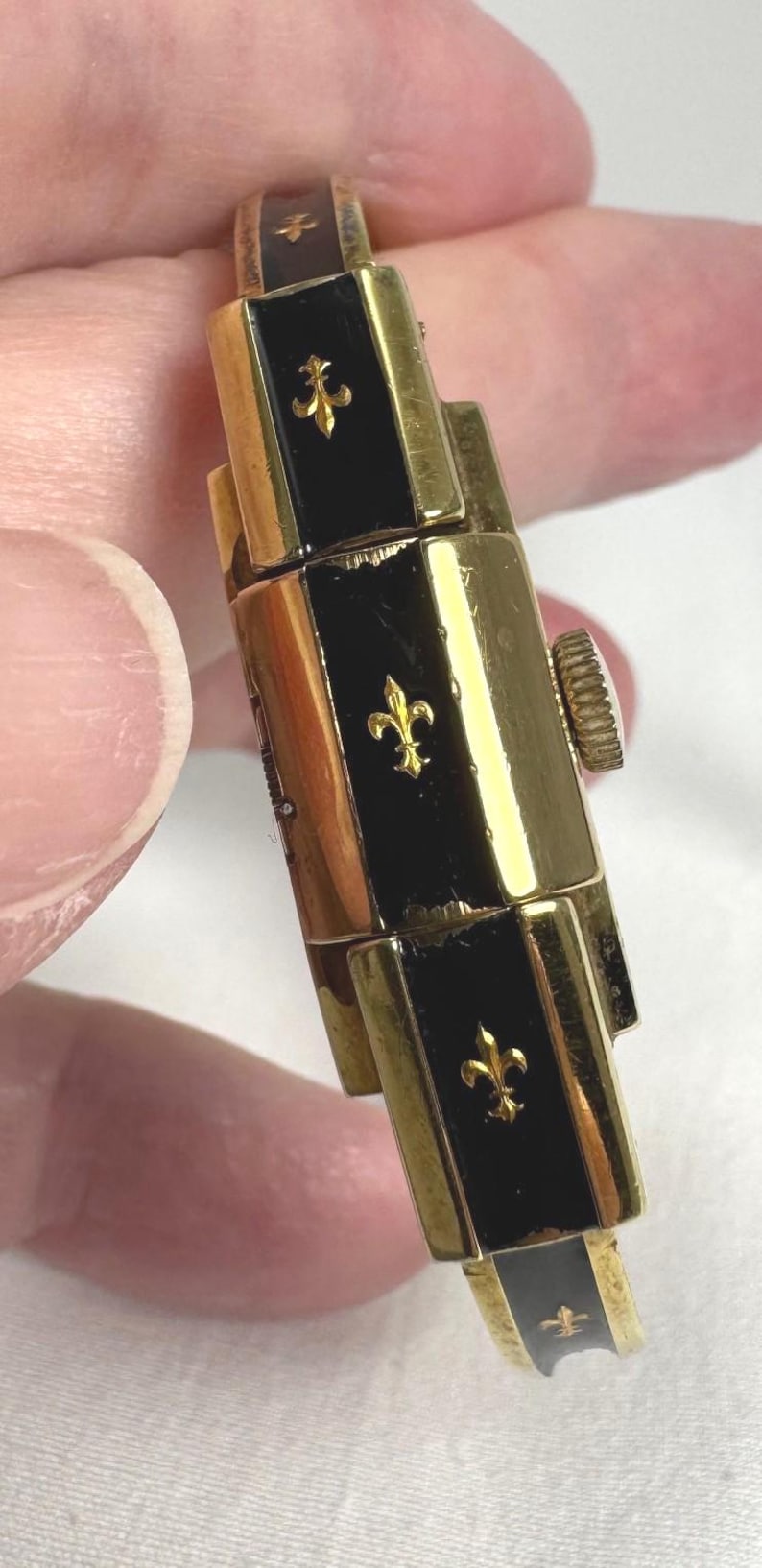 May include: Close-up of a vintage gold-toned watch bracelet with black enamel panels. Each panel features a gold fleur-de-lis emblem. The watch has a textured crown and a classic design.