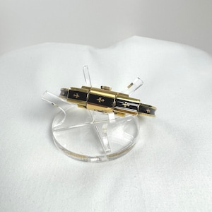May include: A gold-coloured bracelet with black enamel detailing and cross motifs. The bracelet is presented on a clear acrylic stand against a white backdrop. The design of the bracelet incorporates a sequence of rectangular sections.