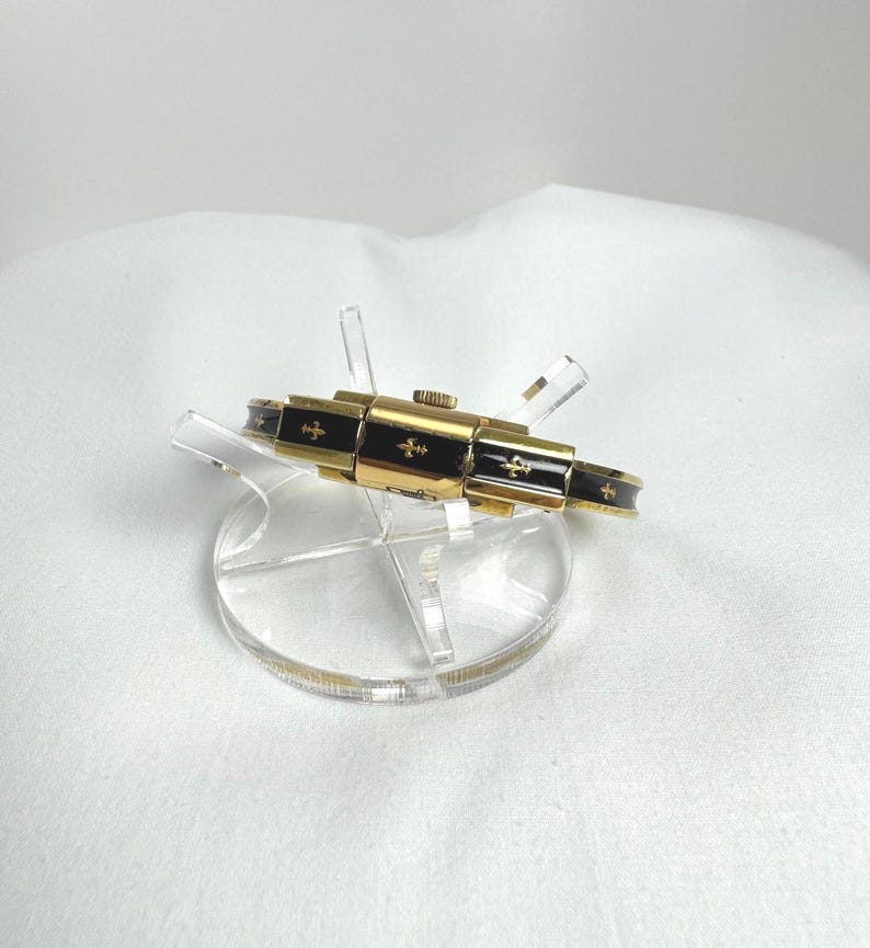 May include: A gold-coloured bracelet with black enamel detailing and cross motifs. The bracelet is presented on a clear acrylic stand against a white backdrop. The design of the bracelet incorporates a sequence of rectangular sections.