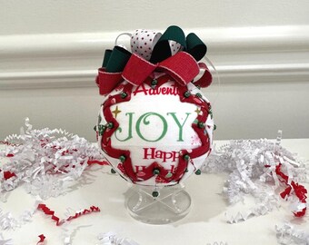 May include: A white Christmas ornament with the word "JOY" in green script, framed by red trim and green accents. The ornament is topped with red, green, and white polka dot ribbons. The words "Advent" and "Happy Holidays" are also visible.