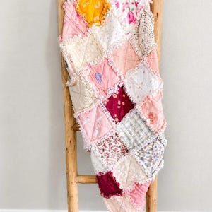 Op de afbeelding: Een handgemaakte patchworkquilt in roze, witte en gele tinten, gedrapeerd over een houten ladder. De quilt heeft verschillende bloemen- en patroonstoffen, met gefranjerde randen. Een gezellig, decoratief item.