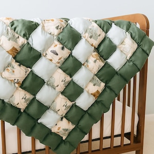 May include: A handmade baby quilt with a forest theme. The quilt features a patchwork design with alternating squares of green, light blue, and patterned fabric with trees and mountains. The quilt is draped over a wooden cot.