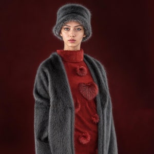 Könnte beinhalten: Eine Frau trägt einen langen, dunkelgrauen Pelzmantel mit einem roten Pulloverkleid, das mit Pelzherzen verziert ist. Sie trägt auch einen grauen Pelzhut.