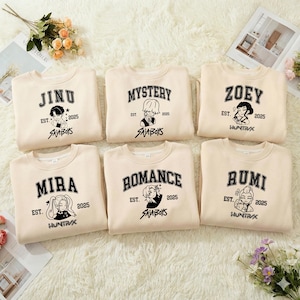 Puede incluir: Seis sudaderas beige de cuello redondo con diseños gráficos negros. Cada sudadera presenta un nombre, el año 2025 y el texto "SAJABOYS" o "HUNTRAX". Los nombres son "JINU", "MYSTERY", "ZOEY", "MIRA", "ROMANCE" y "RUMI".
