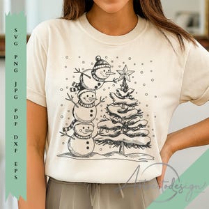 Puede incluir: Camiseta color crema con una ilustración en blanco y negro de una pila de muñecos de nieve y un árbol de Navidad. Los muñecos de nieve llevan sombreros y bufandas. Una estrella corona el árbol. Tema festivo de invierno.