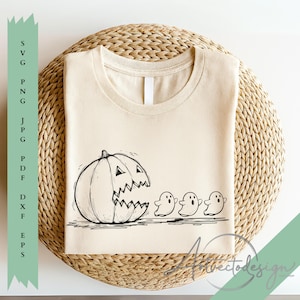 May include: Cream-colored t-shirt featuring a black and white illustration of a jack-o'-lantern with a wide, jagged mouth, chasing three small ghost figures. The shirt is folded on a woven, circular surface, perfect for Halloween.