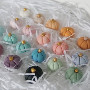 May include: A collection of colorful, miniature pumpkin decorations in various shades of pink, orange, blue, green, and purple, each with a gold-colored stem. The pumpkins are arranged on a bed of white shredded paper.