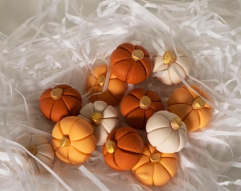 May include: A collection of small, decorative pumpkins in various shades of orange, white, and brown. The pumpkins have a matte finish and are decorated with gold accents. The pumpkins are arranged in a pile on a white background.