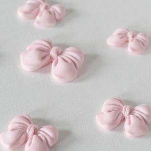 May include: Several small, light pink bow-shaped decorations are scattered on a white surface. The bows have a smooth, matte finish and are evenly spaced, creating a delicate and charming aesthetic. These could be used for crafts or cake decorating.