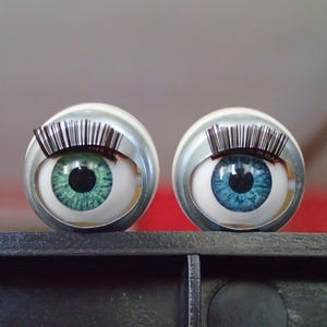 Puede incluir: Dos adornos esféricos con forma de ojos, uno verde azulado y otro azul, ambos con pupilas negras y largas pestañas plateadas. Los ojos están engarzados en una carcasa plateada.