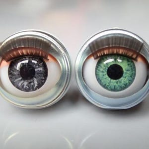 Op de afbeelding: Twee realistische oogcabochons, één met een grijze iris en de andere met een groene iris, gezet in zilverkleurige metalen bezels. De ogen hebben gedetailleerde wimpers en zijn geplaatst tegen een vage achtergrond.