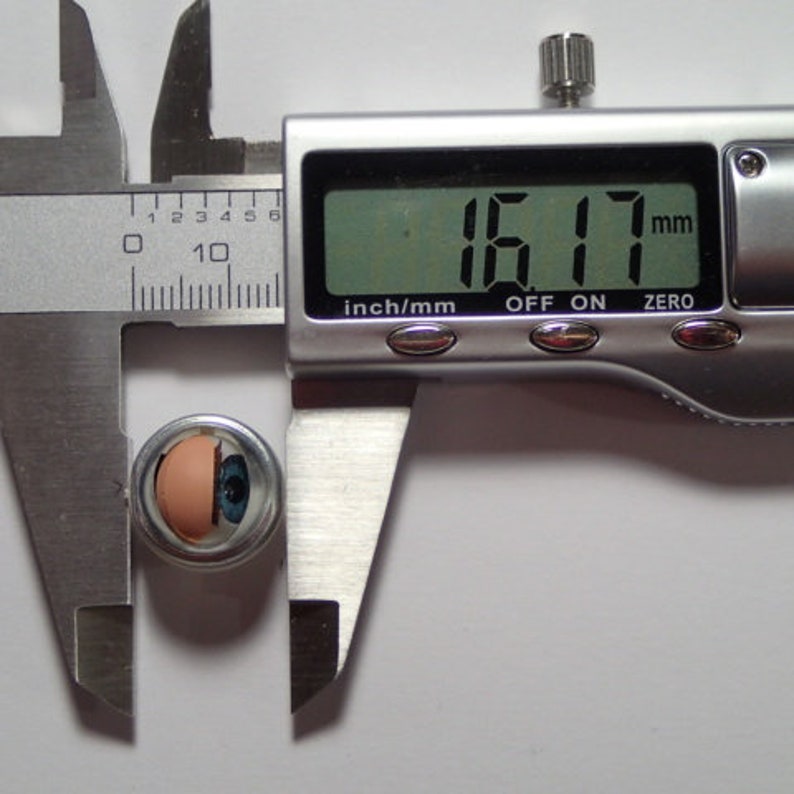 May include: A close-up of a silver caliper measuring a small, round object with a blue and white eye design. The caliper reads 16.17 mm.