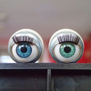 Puede incluir: Dos objetos esféricos con diseños de ojos realistas. Un ojo es azul, el otro verde, ambos con pupilas negras y largas pestañas negras. El fondo está borroso.