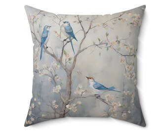 May include: A decorative throw pillow with a watercolor design of blue birds perched on a blooming tree branch with white flowers. The background is a soft blue.