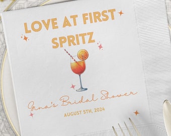 May include: White paper napkin with the text "Love at First Spritz" in orange and pink lettering. A cartoon illustration of a glass of orange juice with a straw and orange slice is above the text. The text "Gina's Bridal Shower" and "August 5th, 2024" is below the illustration.